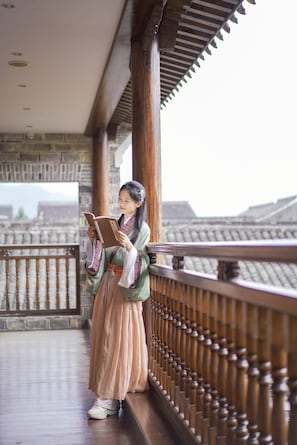 Uma pessoa vestida com traje tradicional lendo um livro numa varanda com guarda de madeira e paredes de pedra ao fundo.