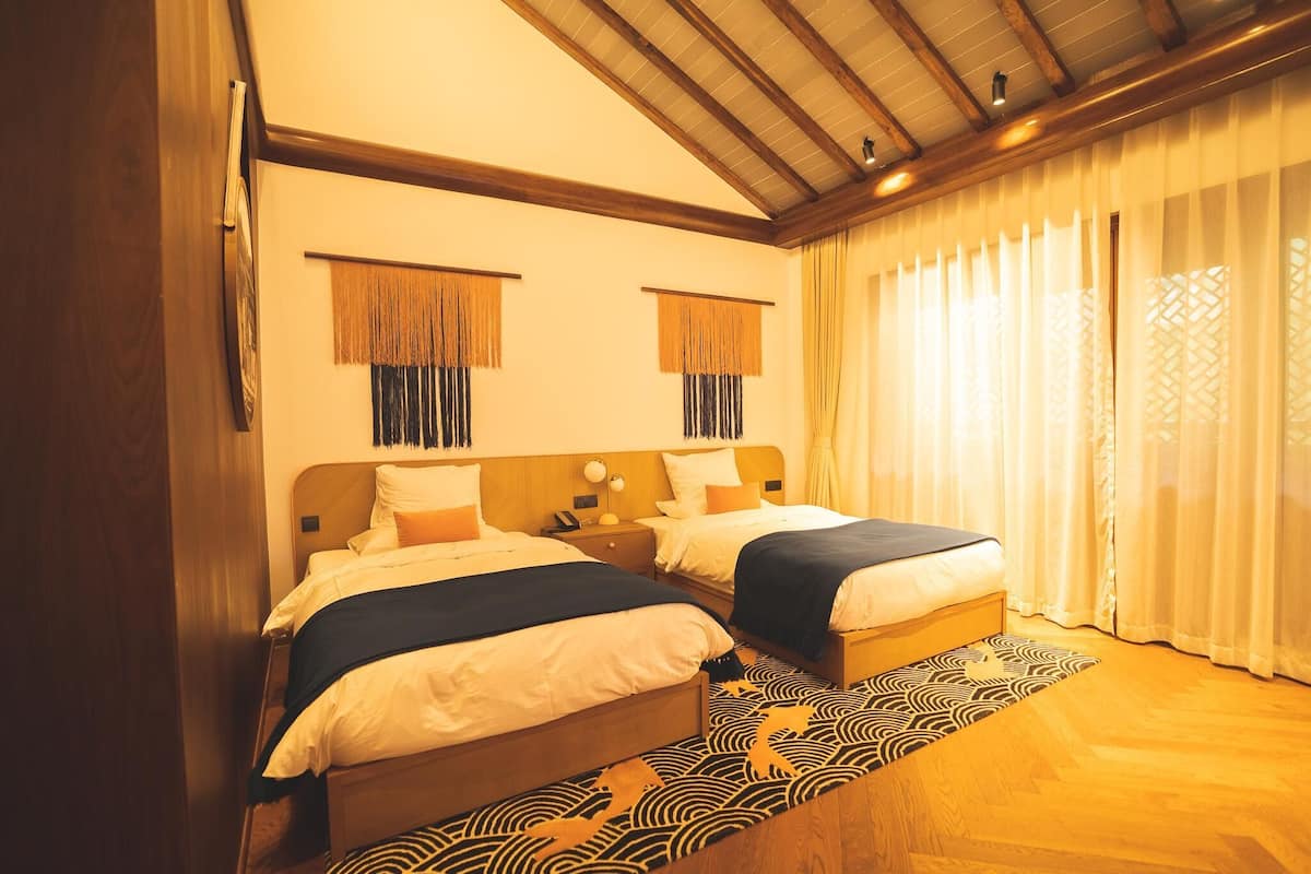 A hotel room with two beds, wooden headboards, a patterned rug, and a large window with sheer curtains.
