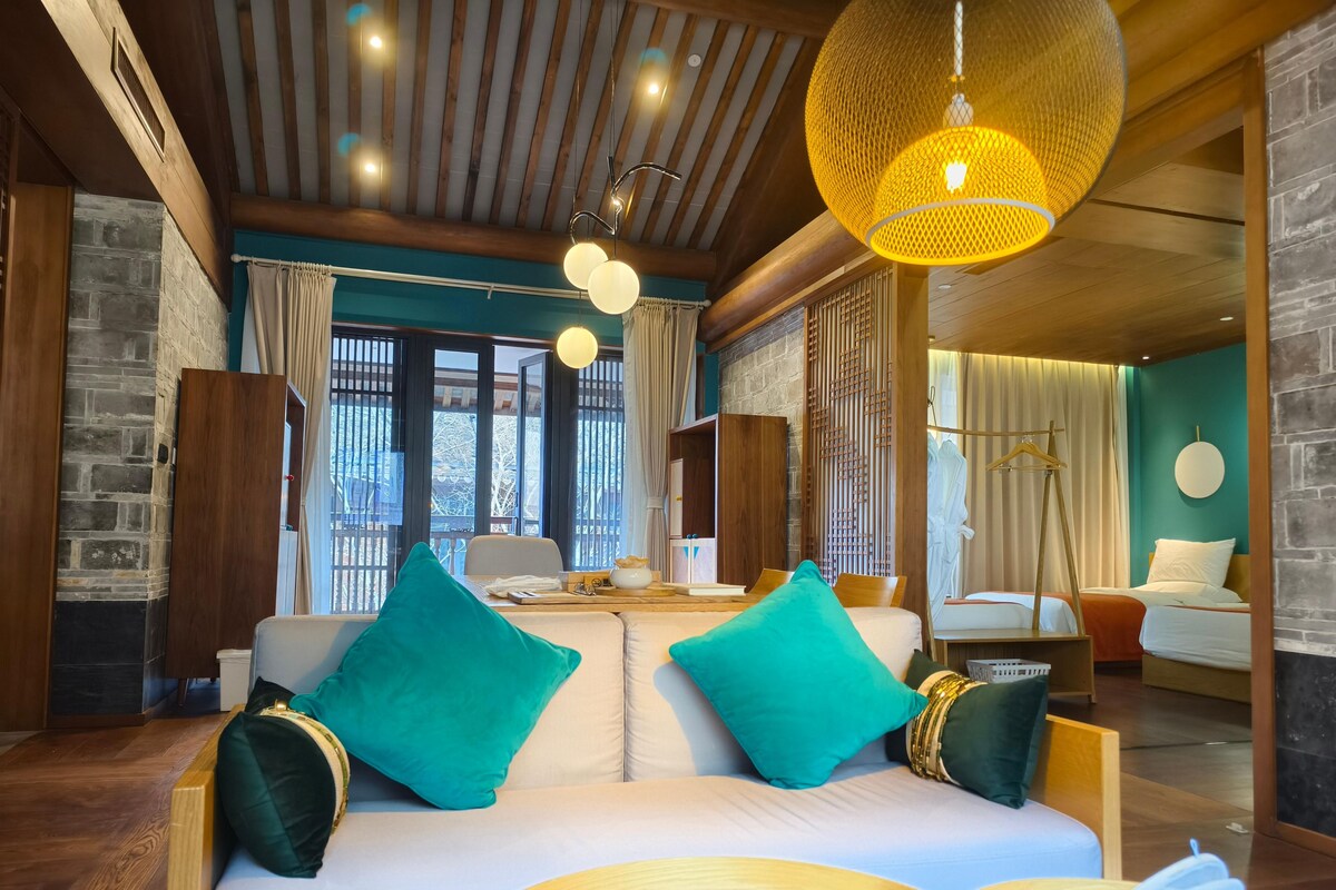 Una habitación de hotel moderna con techo de madera, un ventanal grande, un sofá con cojines color turquesa y una lámpara colgante.
