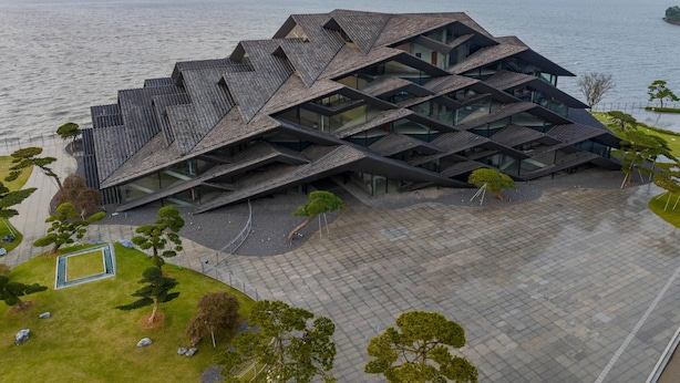 A modern building with a unique geometric roof design, situated by a body of water with mountains in the background. - Blossom Hill Hotel Ningbo Hanling (Ningbo)