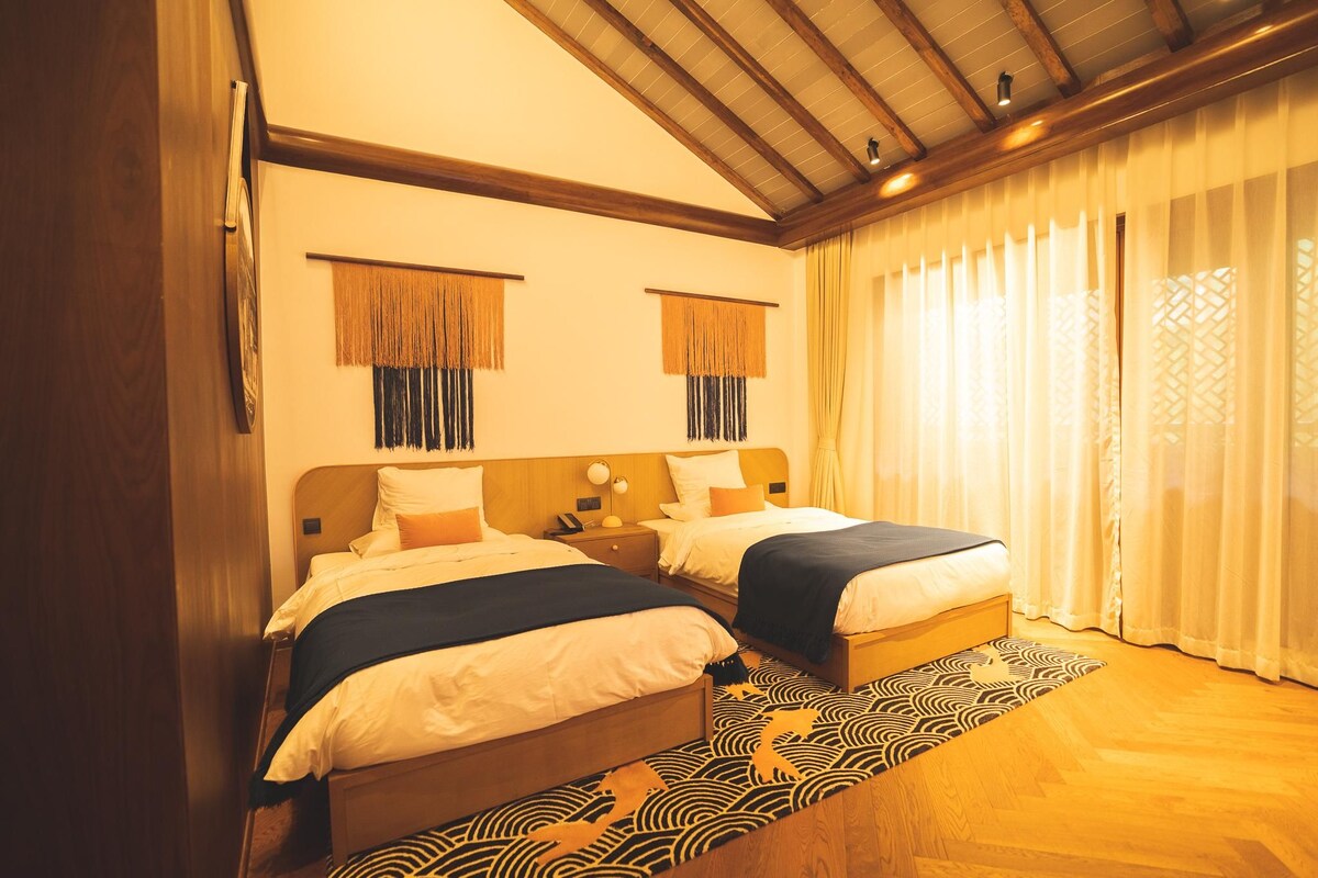 A hotel room with two beds, wooden headboards, a patterned rug, and a large window with curtains.