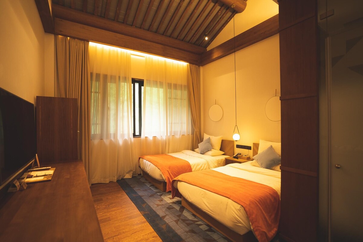 Quarto de hotel com duas camas, uma escrivaninha de madeira, uma mesinha pequena e uma janela com cortinas.