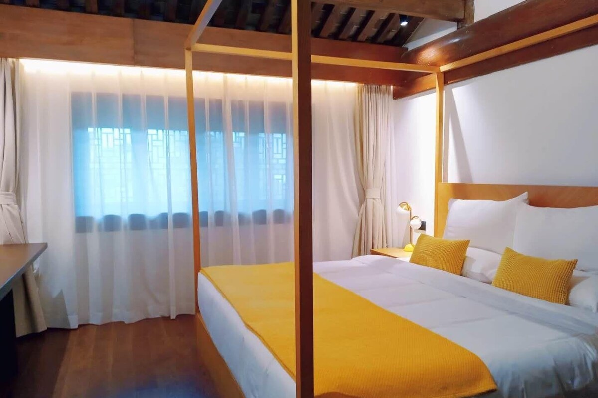 A hotel room with a wooden ceiling, a bed with white and yellow bedding, a desk, and a window with sheer curtains.