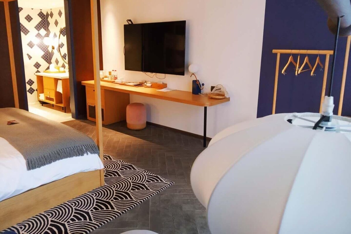 A modern hotel room with two beds, a desk with a flat-screen TV, a wardrobe, and a bathroom visible through an open door.