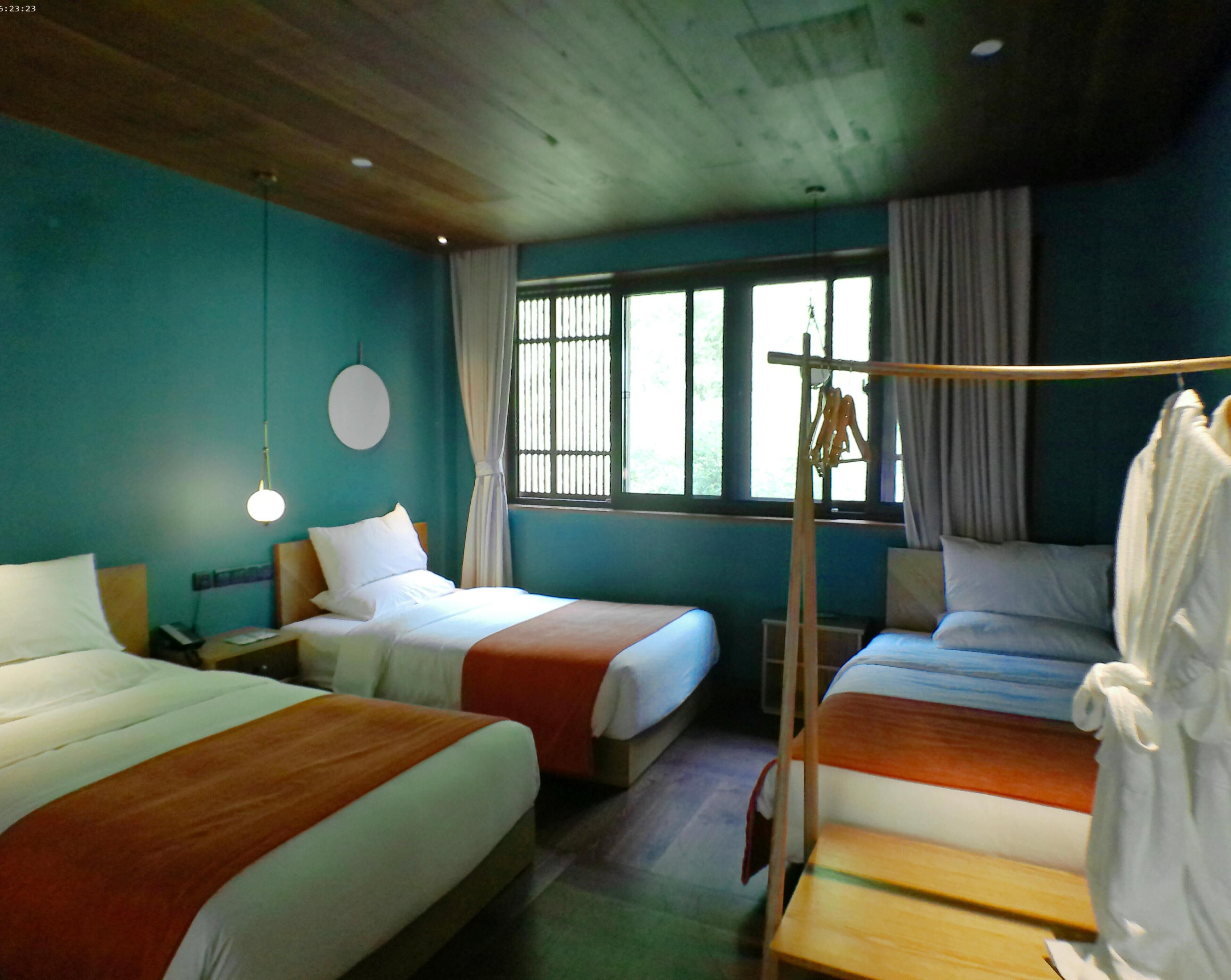 A hotel room with three beds, a wooden ceiling, a large window with curtains, and a hanging rack.