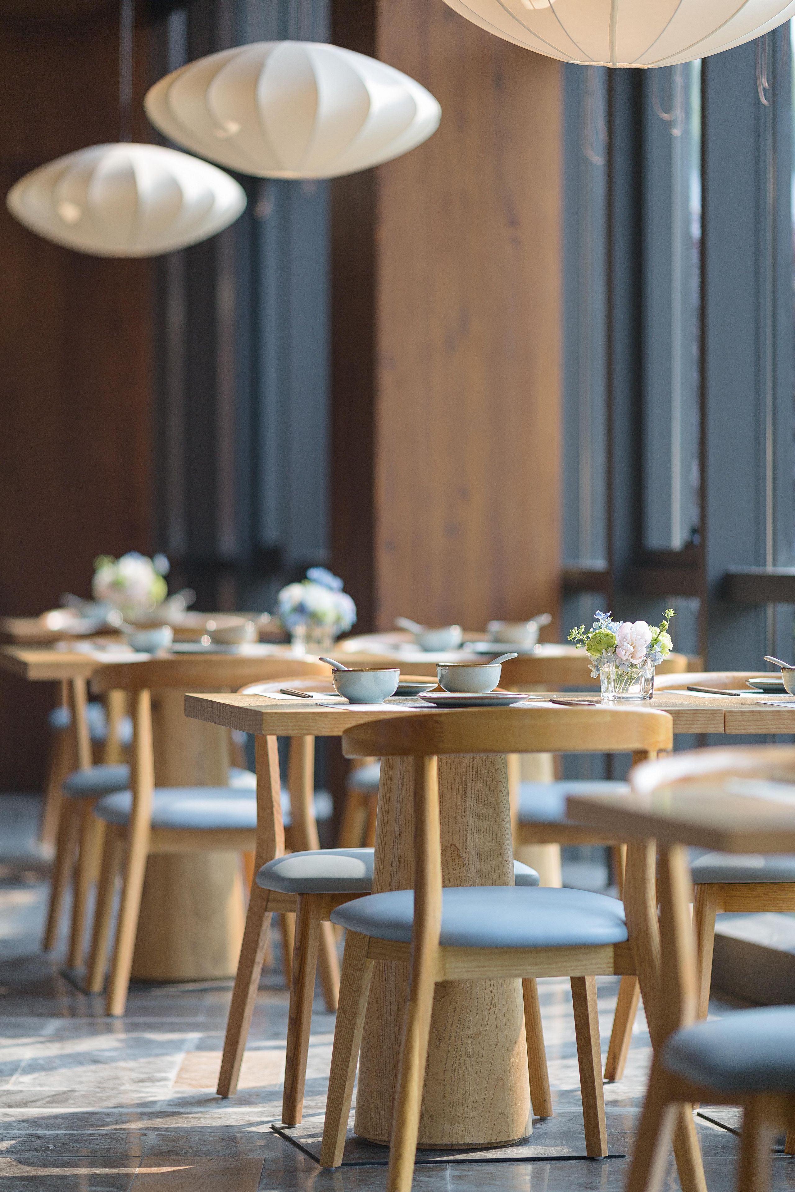 Interior de restaurante con mesas y sillas de madera, lámparas colgantes blancas y centros de mesa con flores.