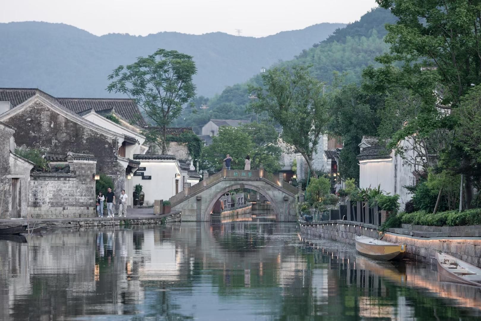 Uma cidade tradicional chinesa à beira da água, com uma ponte de pedra sobre um canal, cercada por vegetação exuberante e edifícios históricos.