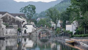 Uma cidade tradicional chinesa à beira da água, com uma ponte de pedra sobre um canal, cercada por vegetação exuberante e edifícios históricos.