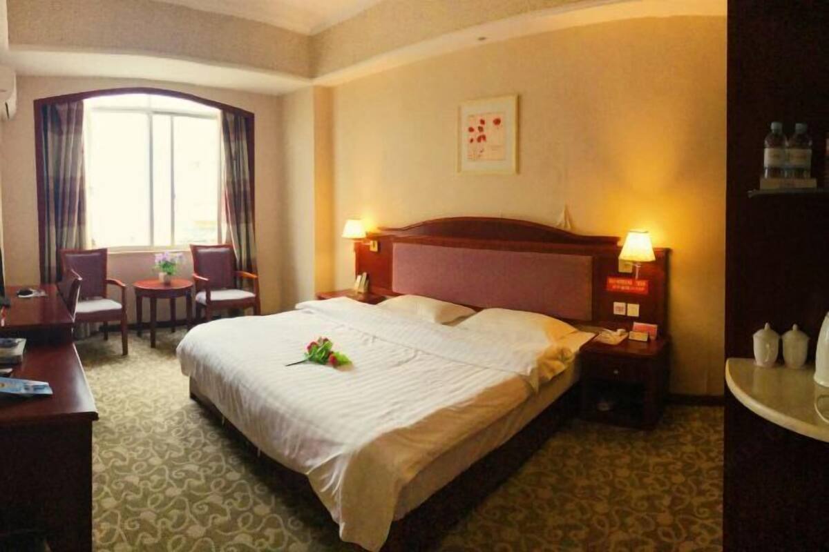 Een hotelkamer met een groot bed, twee bedlampen, een bureau met stoel, een kleine tafel met stoel, en een raam met gordijnen.