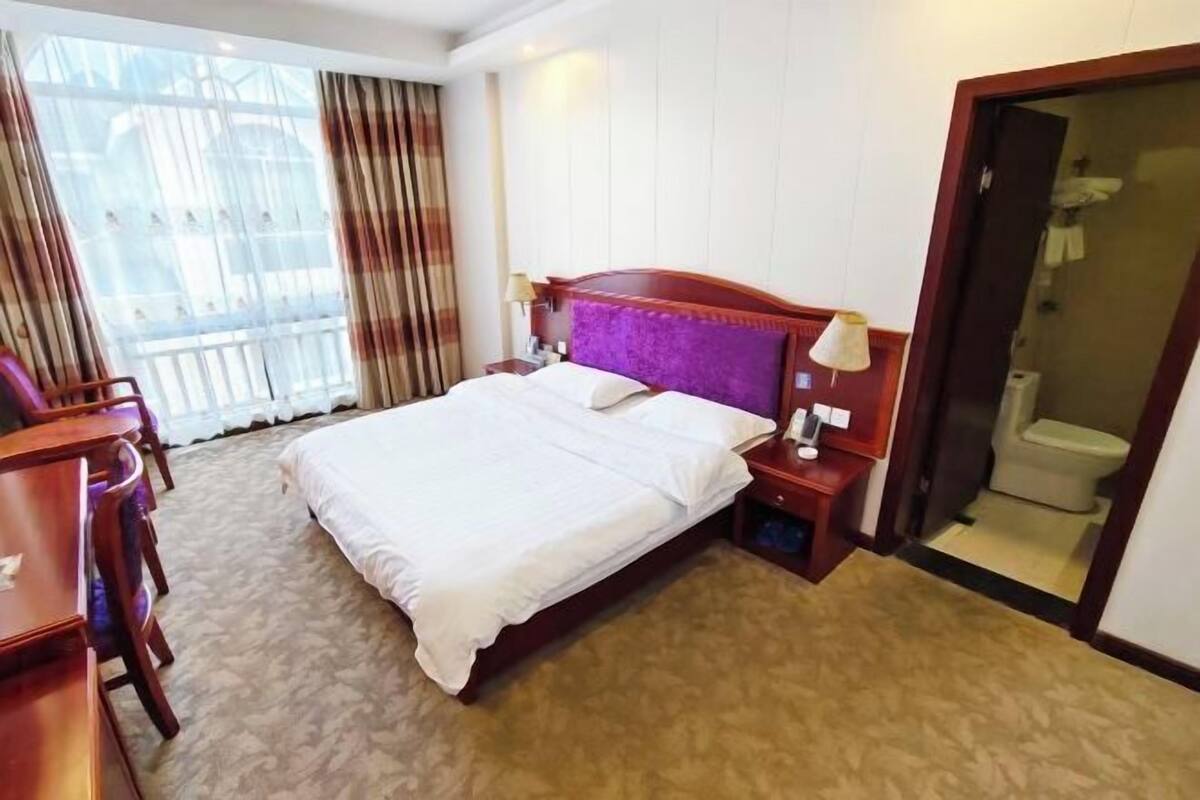 Een hotelkamer met een groot bed, een bureau en een stoel. Er is een raam met gordijnen, een deur naar de badkamer en een nachtkastje met een lamp.