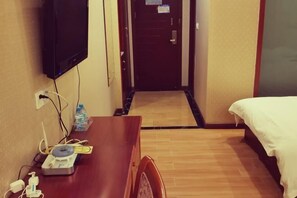 A hotel room with a wooden desk, a flat-screen TV mounted on the wall, a bed with white bedding, and a door leading to another room. - Yongheng Business Hotel (Debao)