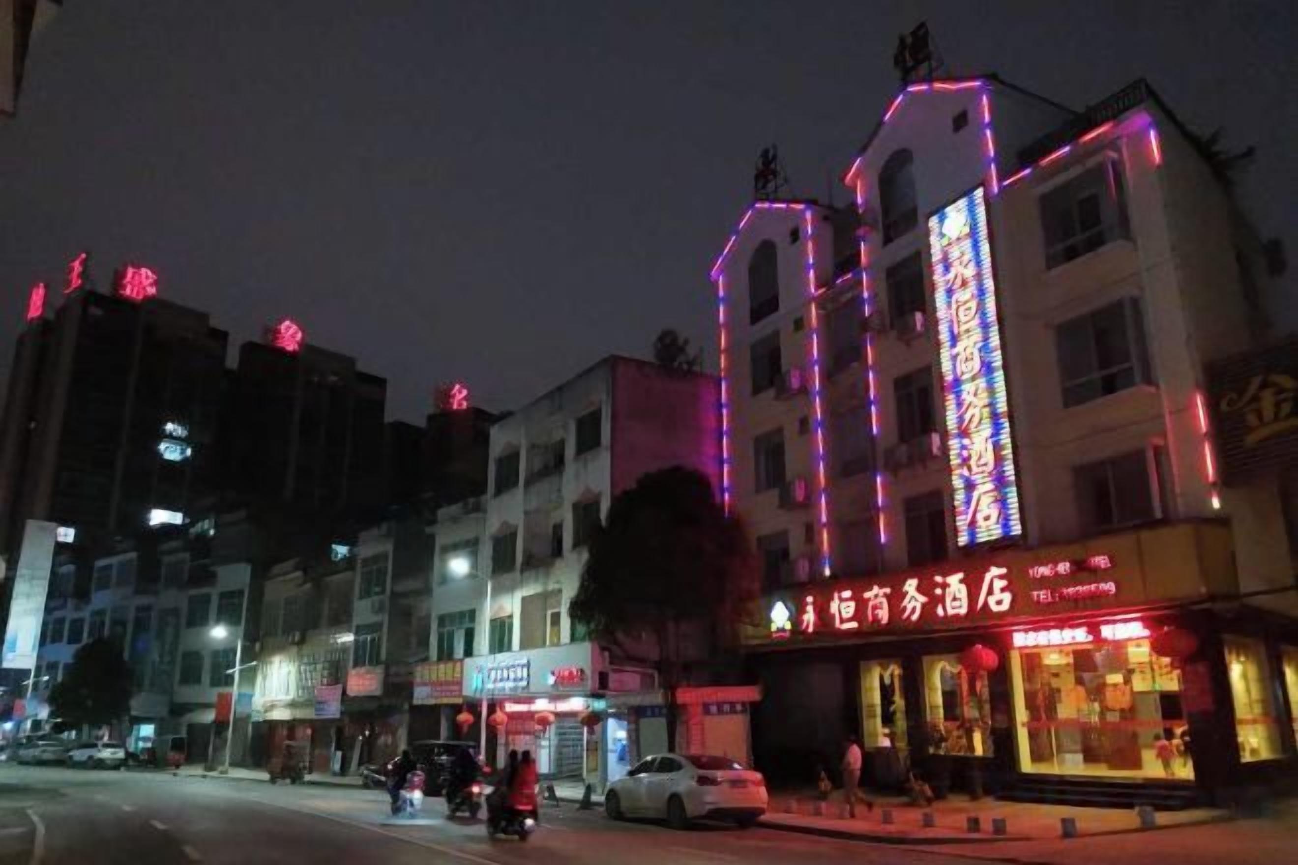 A multi-story building with illuminated signage, a visible restaurant on the ground floor, and a street with parked cars.
