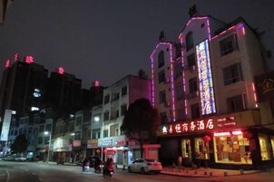 Un edificio de varios pisos con letreros iluminados, un restaurante visible en la planta baja, y una calle con automóviles estacionados.