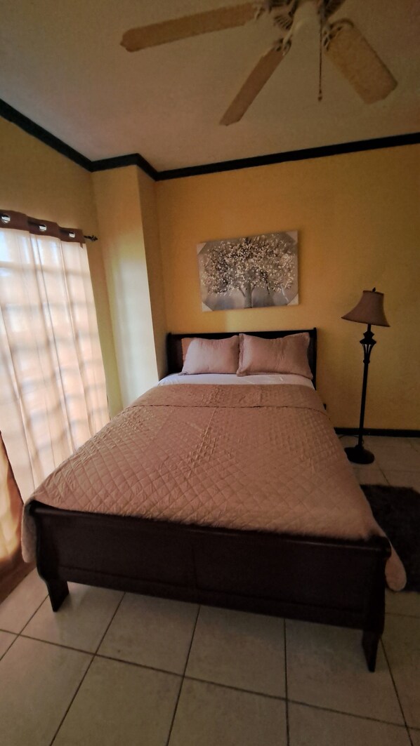 A bedroom with a bed, a ceiling fan, a lamp, and a window with curtains. - The Duplex (Bonne Terre)