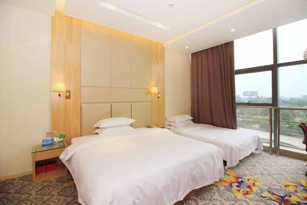 A hotel room with two beds, a nightstand, a window with curtains, and a view of the city.
