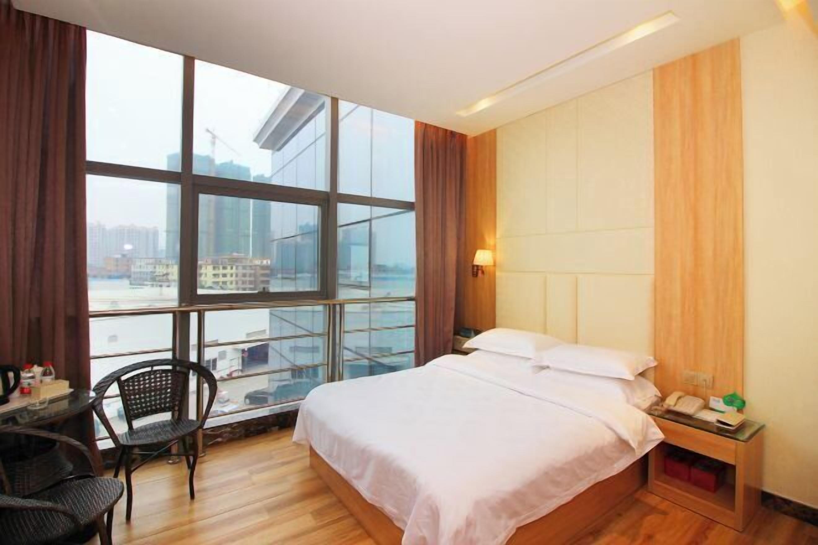 A hotel room with a large window, a bed, a desk with a chair, and a nightstand with a lamp.