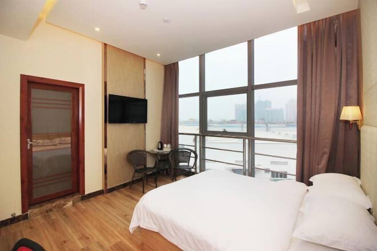 A hotel room with a bed, a TV, a small table with chairs, and a view of the city through large windows.