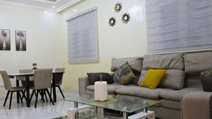 A living room with a glass coffee table, a sofa with cushions, a dining table with chairs, and wall art.