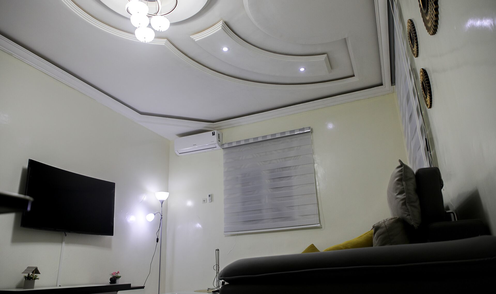 A room with a flat-screen TV mounted on the wall, a ceiling with recessed lighting, a window with blinds, and a sofa with cushions.