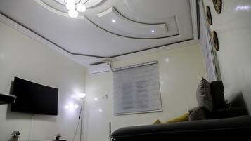 A room with a flat-screen TV mounted on the wall, a ceiling with recessed lighting, a window with blinds, and a sofa with cushions.