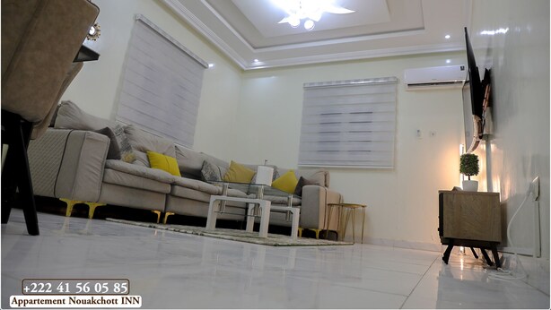A living room with a sofa, coffee table, and a TV mounted on the wall. There is a ceiling light fixture and an air conditioning unit.