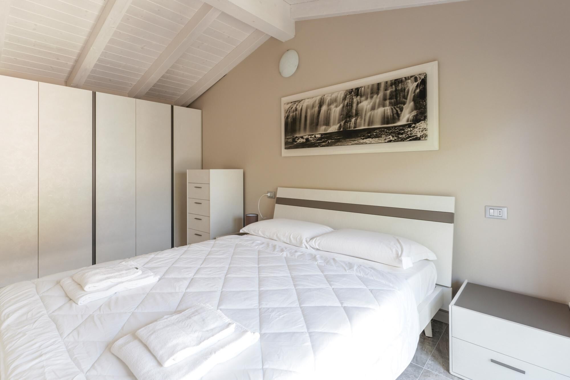 A neatly made bed with white bedding and a folded towel, a bedside table, a wardrobe, and a framed photograph on the wall.