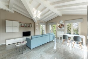 A spacious living area with a blue sofa, a dining table with chairs, a TV, and a white cabinet with shelves.