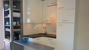 Eine moderne Küche mit weißen Schränken, einer schwarzen Arbeitsplatte und einem Spülbecken. Zwei weiße Hocker und eine Schüssel mit Obst stehen auf der Arbeitsfläche.