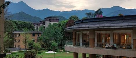 A modern hotel with a curved balcony, lush greenery, and mountains in the background.