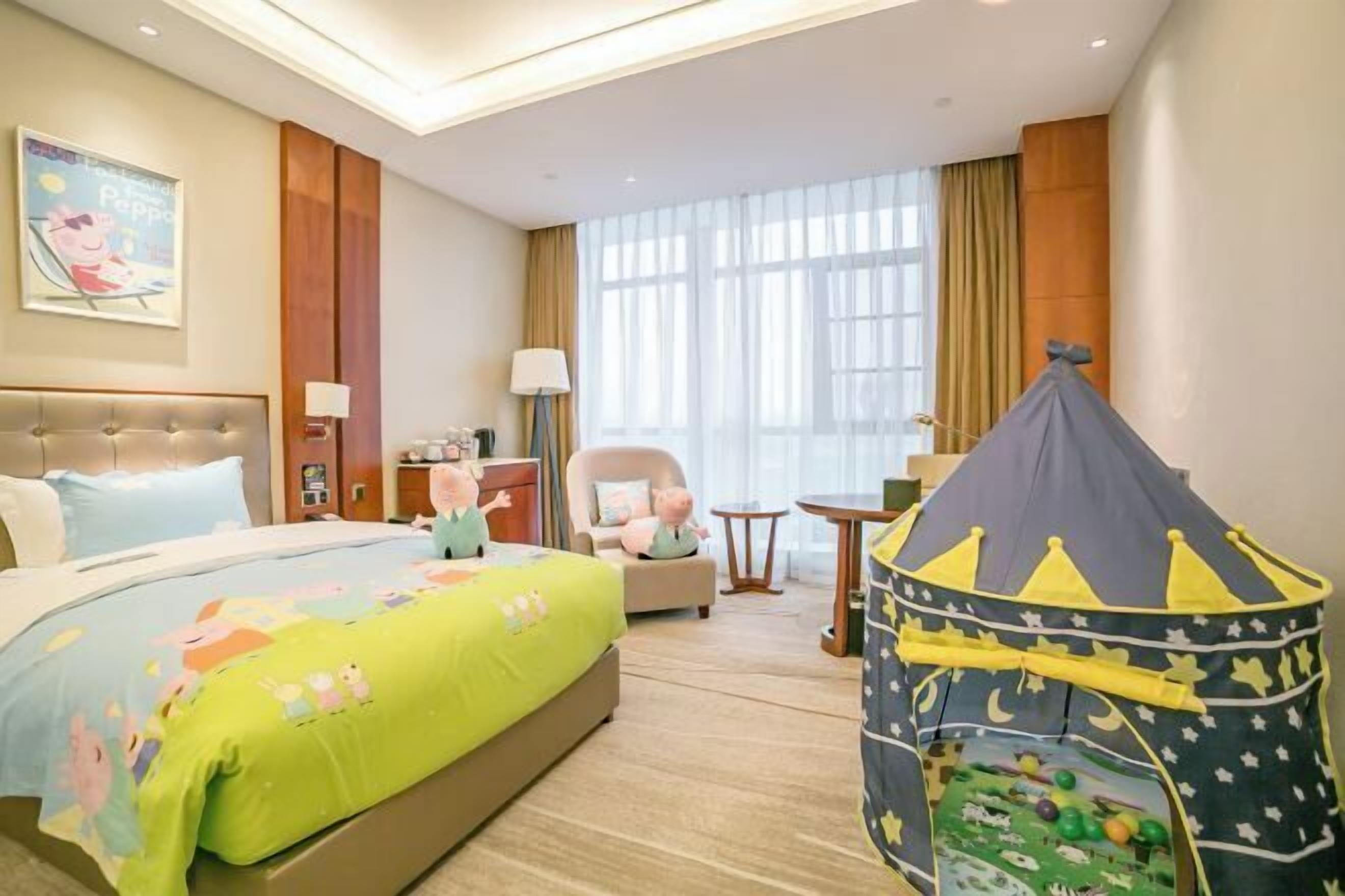 Une chambre d’hôtel avec un lit, une chaise, une petite table, une lampe, une fenêtre avec des rideaux et une tente de jeu.