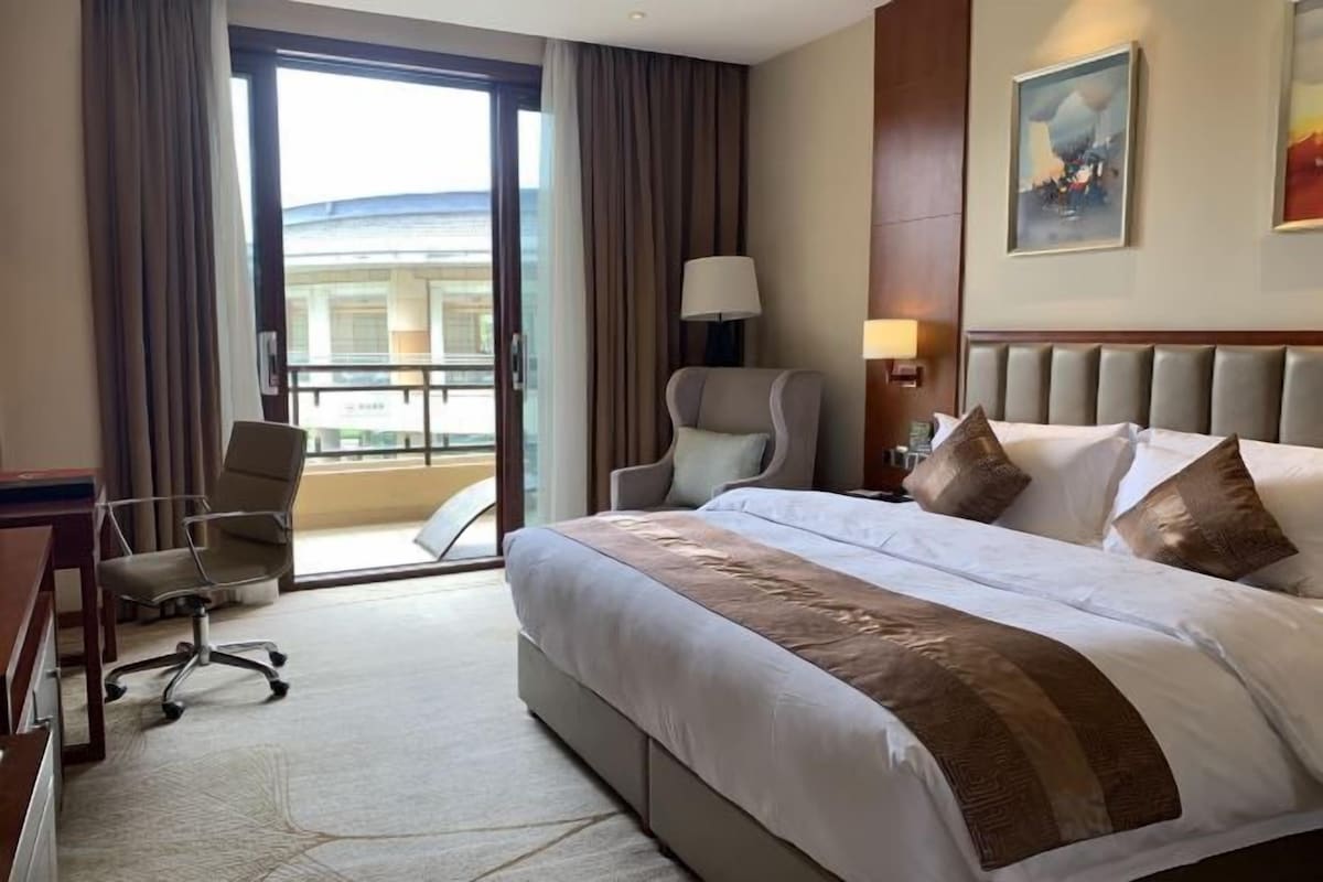 A hotel room with a large bed, a desk, a chair, a lamp, and a view of the outside.