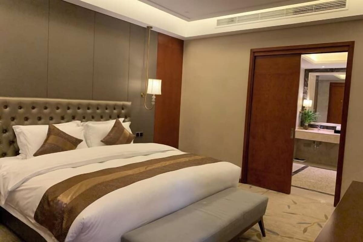 A hotel room with a large bed, a bedside table, a bench, a mirror, and a door leading to a bathroom.