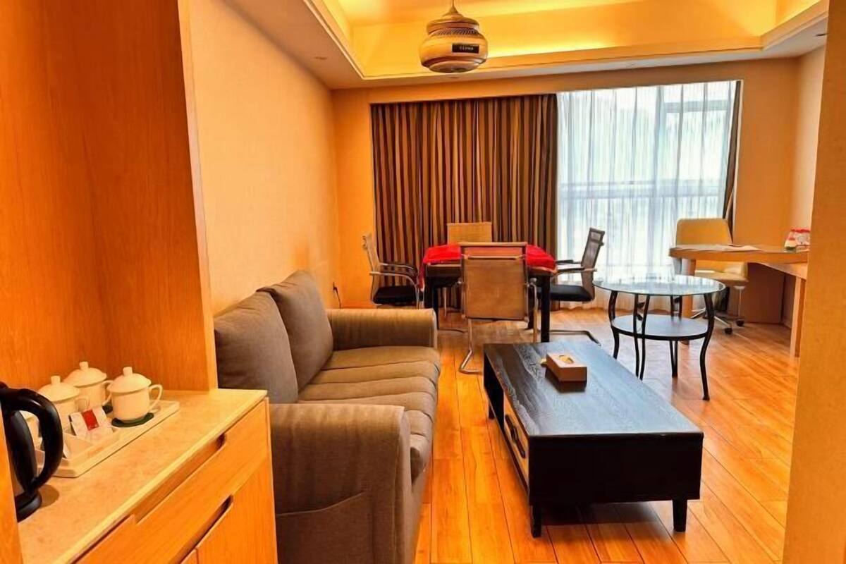 Une chambre d’hôtel compacte comprenant un canapé, une petite table à manger, une table basse et un bureau avec une chaise. On y trouve une fenêtre avec des rideaux, un luminaire au plafond et une petite table avec une bouilloire et des tasses.