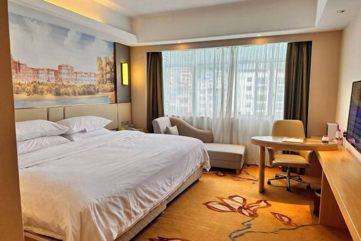 Une chambre d’hôtel avec un grand lit, un bureau, une chaise et une vue sur le paysage urbain, visible à travers de fines rideaux.