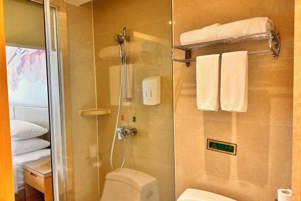 Baño de hotel con ducha de vidrio, inodoro, toallero con toallas y un cartel en la pared.