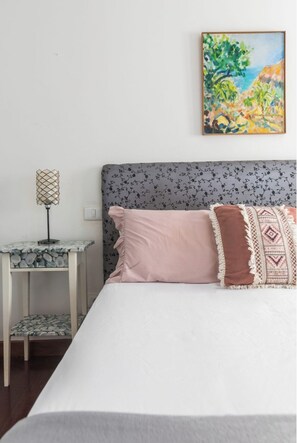 Un dormitorio con cama, mesita de noche, lámpara y un cuadro en la pared.