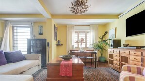 Uma sala de estar com sofá, mesa de centro, mesa de jantar com cadeiras, televisão e uma planta.