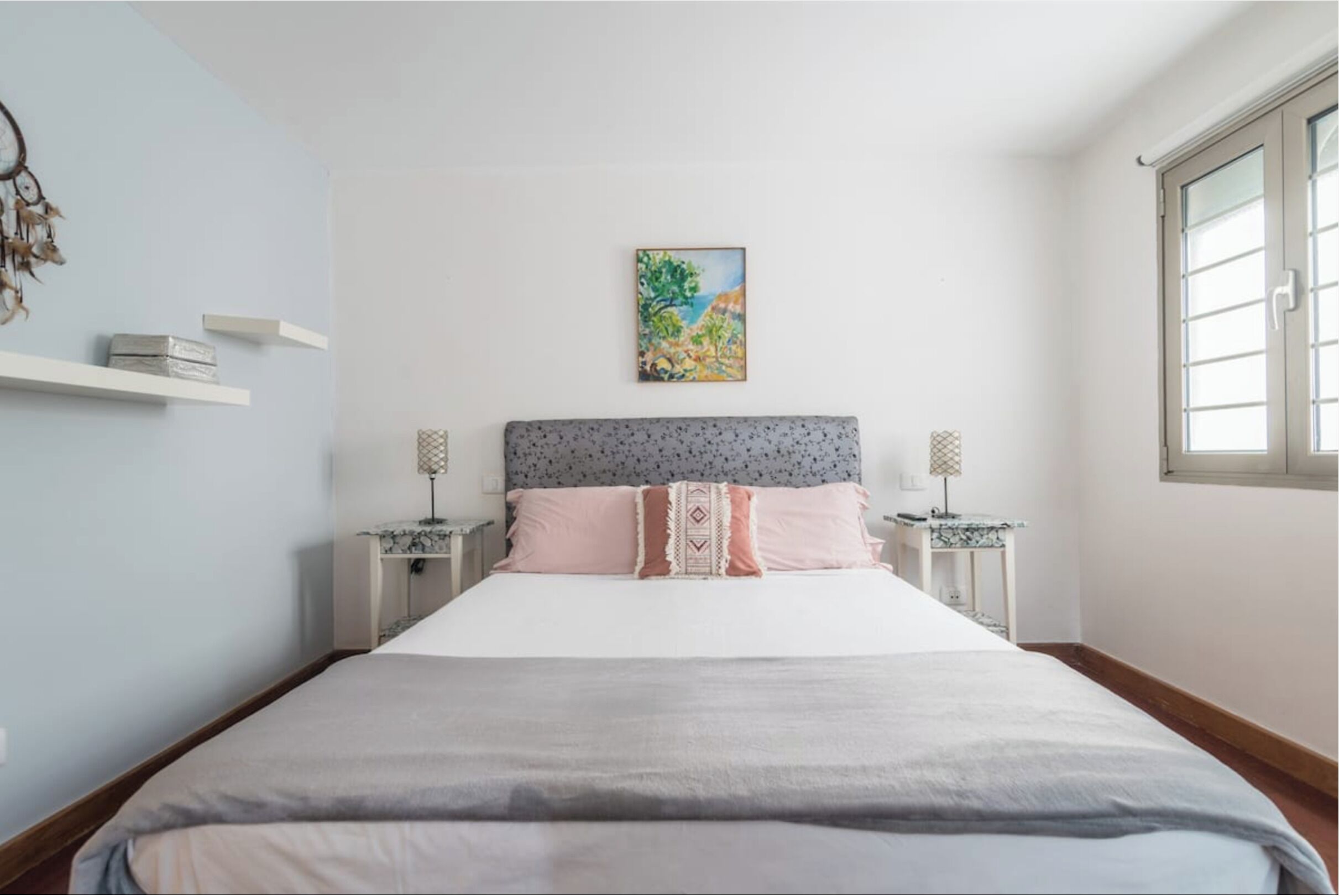 Un dormitorio con cama, dos mesitas de noche, una mesita auxiliar, un estante en la pared, un cuadro y una ventana.