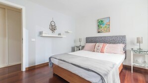Um quarto com piso de madeira, uma cama com cabeceira cinza, um sonho capturado, um quadro e uma mesa de cabeceira com abajur.
