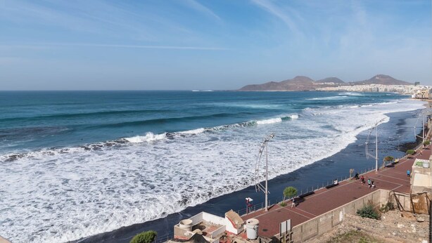 Vista costeira com praia, ondas e uma cadeia montanhosa ao longe.
