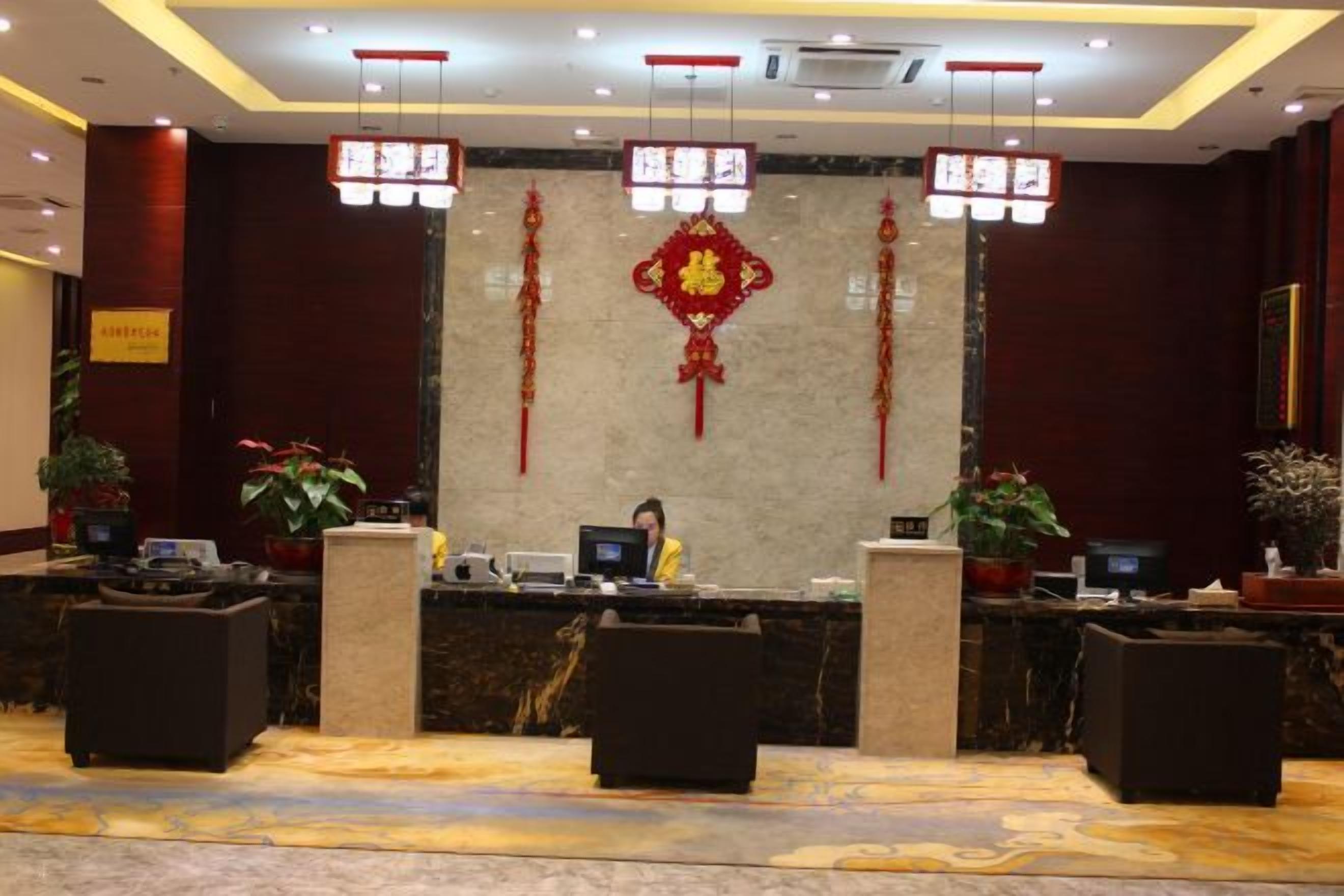Zona de recepción de un hotel con mostrador de mármol, dos recepcionistas y una pared decorativa con un emblema rojo.