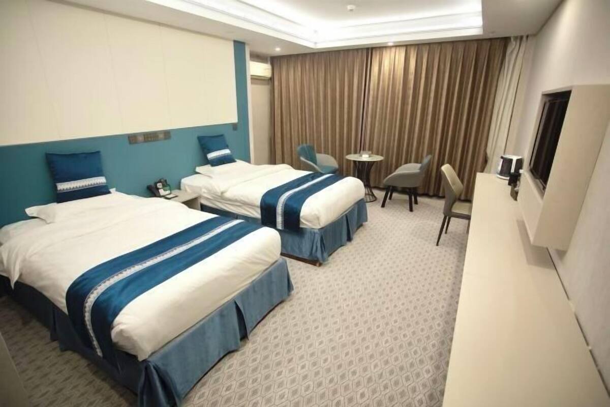 Quarto de hotel com duas camas, uma escrivaninha, uma cadeira, televisão e janela com cortinas.
