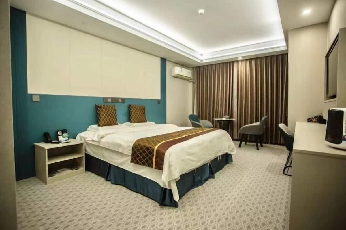Quarto de hotel com cama, escrivaninha, cadeira e uma janela grande.