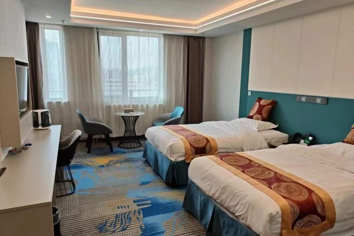 Quarto de hotel com duas camas, uma escrivaninha, uma cadeira, uma mesinha e uma janela com cortinas.