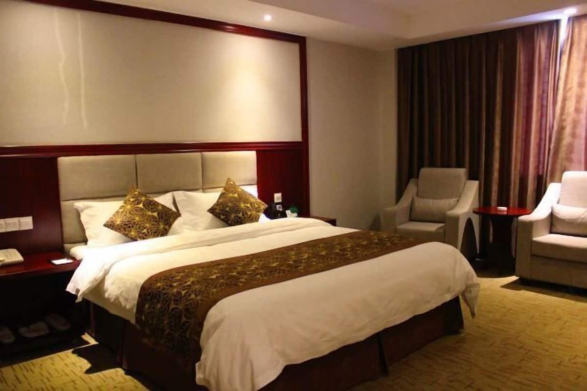 Quarto de hotel com uma cama grande, duas poltronas, uma mesa pequena e uma mesinha de cabeceira com um telefone e uma garrafa.