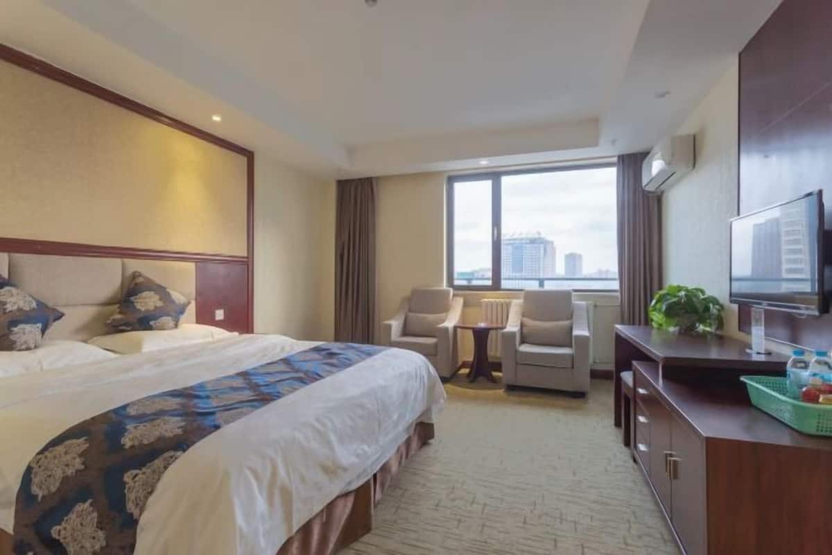 Quarto de hotel com uma cama grande, duas poltronas, uma mesinha, televisão e vista para a cidade.