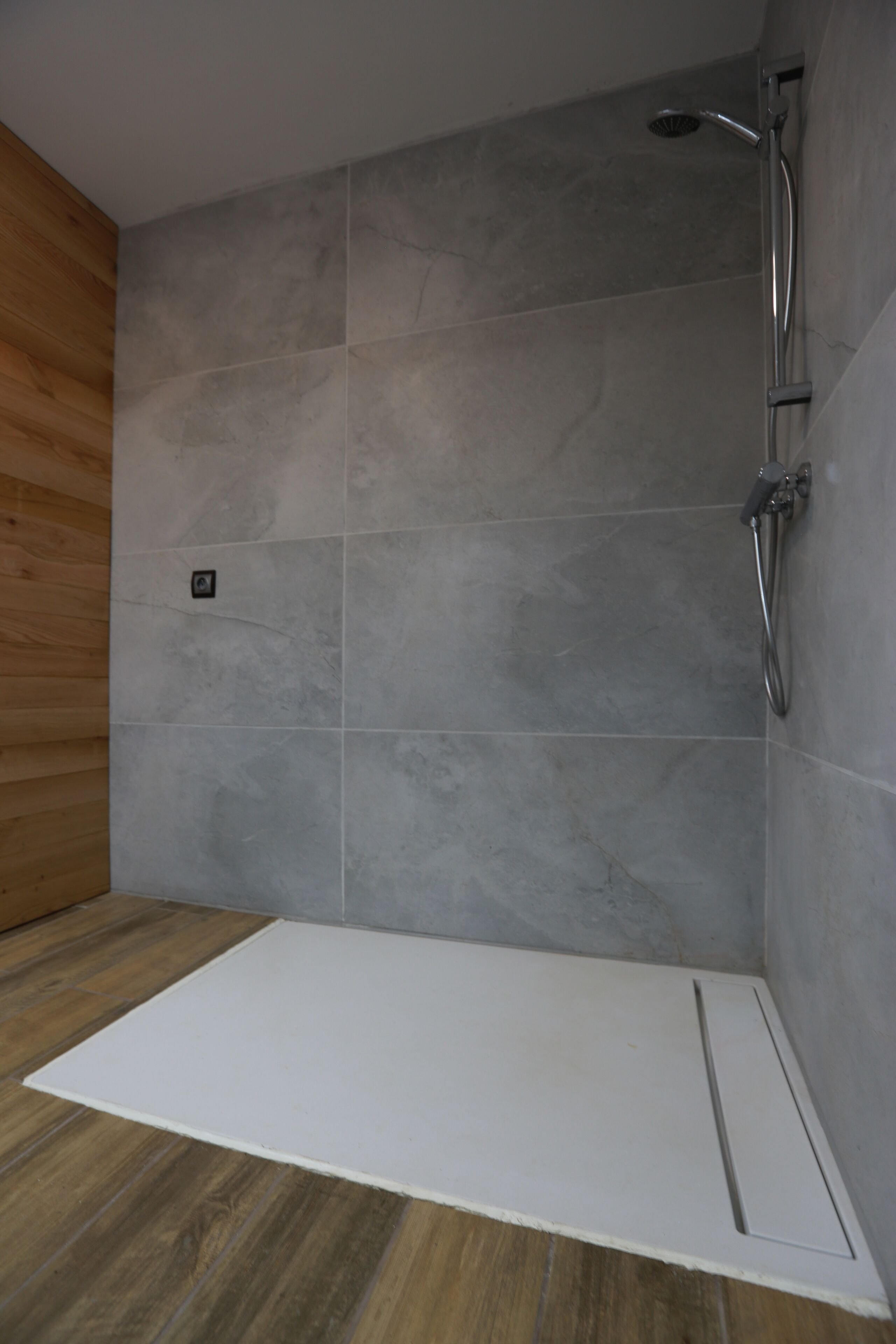 A shower area with grey tiles, a wooden panel wall, and a white floor mat.
