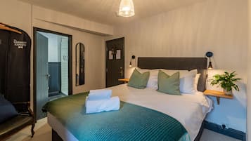 Una cama bien hecha con un edredón verde, una lámpara de noche y una planta en una mesita.