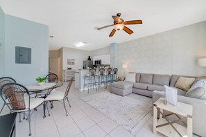 A modern living room with a grey sofa, a dining table with chairs, a kitchen area with a fridge and microwave, and a ceiling fan.