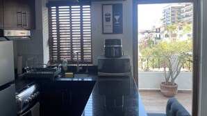 A modern kitchen with a central island, a stove, a sink, and a coffee maker. There is a window with blinds, a potted plant on the balcony, and a view of a residential area. - Le Petit Coeur • Beautiful Ocean View Romantic Zone (Puerto Vallarta)